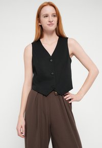 Črna telovnik z V-izrezom, z tremi gumbi in skrajšanim, krojenim dizajnom. Nošeno preko hlač z visokim pasom v rjavi barvi. Gladek videz.