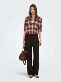 Jeune femme portant une chemise à carreaux rouge, un pantalon noir à jambes larges, des chaussures couleur sable, tenant un sac à main en cuir marron, debout devant un fond blanc.