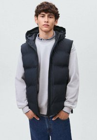 Črna oblazinjena brezrokavnik s kapuco, sprednjim zadrgo in dvema stranskima žepoma, nosi se čez svetlo sivo majico s kapuco in modre kavbojke. Gladek sintetični material.