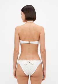 Bela bikinka iz teksturiranega blaga, ki vključuje bandeau top z zlatim sponko in spodnji del z nagubanim detajlom. Valoviti robovi.