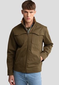 Olijfgroene jas met een ritssluiting aan de voorkant, twee borstzakken en een bruine kraag. Gemaakt van gladde stof met een gestructureerde pasvorm, gecombineerd met lichtblauwe jeans.