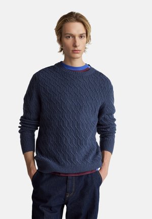 Marineblauer Strickpullover mit strukturierter Musterung, Rundhalsausschnitt und gerippten Bündchen. Über einem mehrlagigen Hemd getragen und kombiniert mit dunklen Jeans.