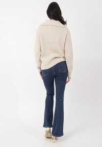Beige gerippter Pullover mit weitem Kragen, kombiniert mit weit geschnittenen dunkelblauen Jeans und beigen hohen Schuhen, von hinten betrachtet.
