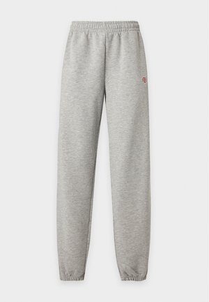 Jeune femme debout portant une chemise blanche à manches courtes, un pantalon de jogging gris avec un petit logo rouge, et des chaussures plates noires avec des brides.