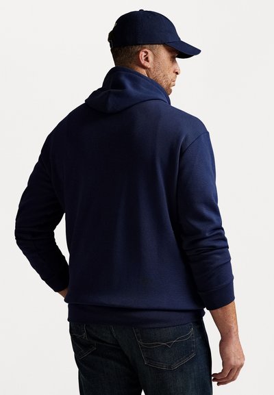 Sweatshirt azul-marinho com capuz e mangas longas, feito de tecido texturizado, combinado com jeans escuros e um boné de basebol combinante.