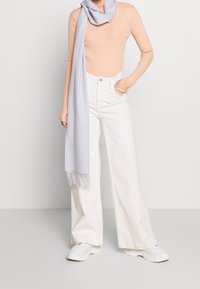 Top lungo a maniche lunghe a coste color pesca, pantaloni bianchi a gamba larga, sciarpa grigia chiara con frange e scarpe sportive bianche, su uno sfondo neutro.