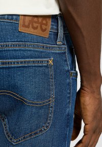 Jeans de mezclilla azul con un parche de cuero que muestra "Lee". Presenta costuras en contraste y un clásico detalle en los bolsillos traseros.