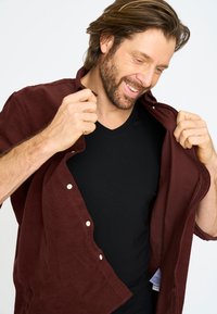 Bordeaux shirt van een zachte, gestructureerde stof, met knopen aan de voorkant en een relaxte pasvorm, gedragen over een zwart V-hals shirt.