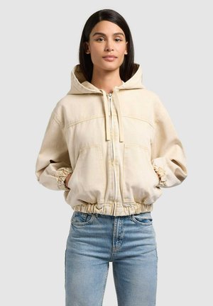 Kvinde med glat sort hår iført en cremefarvet hættetrøje med lynlås og elastiske manchetter samt lyseblå jeans, stående foran en ensfarvet baggrund.