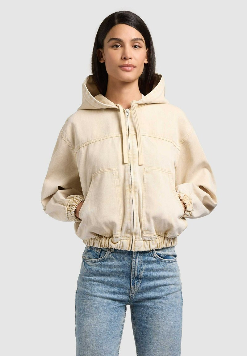 Femme aux cheveux noirs et raides portant un sweat à capuche crème zippé avec des poignets élastiques et un jean bleu clair, debout devant un fond uni.