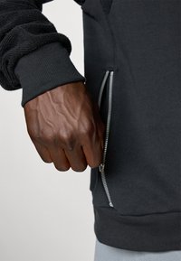 Dark gray sweatshirt with textured sleeves and a silver zipper. Close-up of a clenched hand near the zippered pocket. Smooth fabric finish.