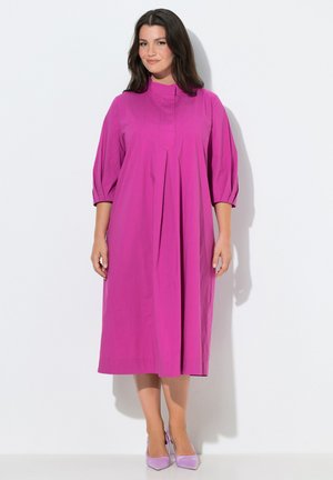 Kvinde med mørkt hår iført en knaldrød midi-kjole med trekvartærmer og lavendelspidse hæle, stående mod en hvid baggrund.