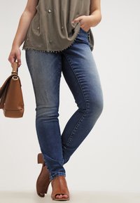 Siniset farkut, joissa on kapeat lahkeet, haalistuneet yksityiskohdat ja sileä pinta, yhdistettynä väljäntuntuiseen oliivinvihreään paitaan ja ruskeaan käsilaukkuun. Ruskeat korkeat sandaalit.