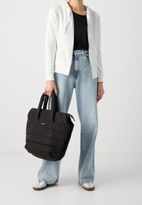 Borsa tote nera imbottita con manici corti, design trapuntato e un piccolo etichetta del marchio, abbinata a jeans flare chiari e una felpa bianca.