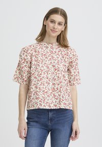 Blouse à manches courtes à motif floral en crème avec des motifs rouges et bleus, dotée d'un col rond et d'une fermeture à boutons, associée à un jean en denim bleu.