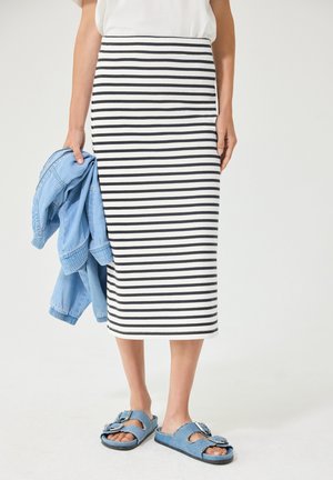 Kvinde iført sort-hvid, horisontalt striped midi nederdel og blå sandaler, holder en lyseblå denimjakke i venstre hånd.