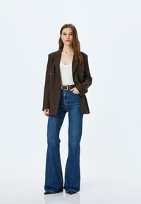 Blazer marrone doppiopetto sopra una maglietta bianca, abbinato a jeans in denim blu a zampa di elefante. Dettaglio della cintura notevole e scarpe nere eleganti.