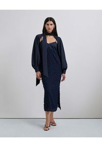 Vestido de satín azul marino con lunares texturizados, que presenta un escote suave y mangas abullonadas, acompañado de sandalias negras con tiras.