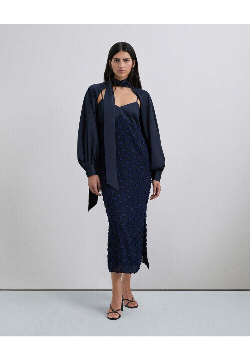 Vestido de satín azul marino con lunares texturizados, que presenta un escote suave y mangas abullonadas, acompañado de sandalias negras con tiras.