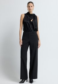 Zwarte satijnen mouwloze top met gedrapeerde halslijn, gecombineerd met zwarte high-waisted broek en zwarte puntige hakken. Strakke lijnen en een getailleerde silhouet.