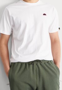 Hvid bomulds t-shirt med et lille pink og sort logo på venstre bryst. Par med grønne sweatpants med snoretræk. Blød tekstur.