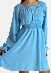 Ljusblå klänning med långa ärmar, resår i midjan och knappdetalj vid halsringningen. Mjukt, texturerat tyg med en flytande fåll.