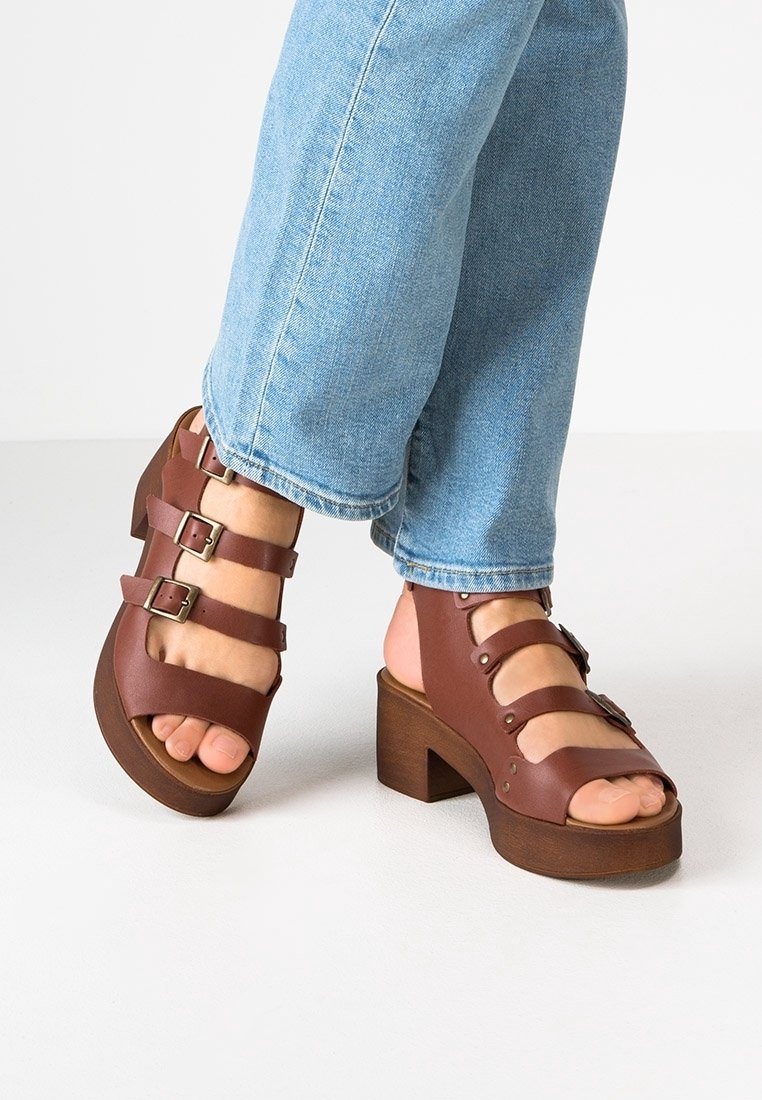 Pieds portant des sandales compensées en cuir marron avec trois boucles chacune, assorties à un jean en denim bleu clair sur un fond uni.