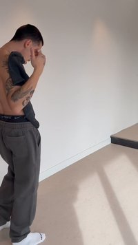 Osoba bez koszulki z tatuażami trzyma czarną koszulę, patrząc w dół, stojąc obok białej ściany i beżowej podłogi z ciemnym stopniem.