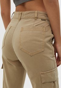 Beige cargobroek met een hoge taille, grote zijzakken en versterkte stiksels. De stof lijkt stevig met een gladde textuur.