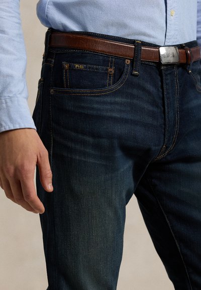 Bărbat purtând blugi albastru închis cu o curea maro din piele și o cămașă bleu deschis cu nasturi, mâna relaxată pe lângă corp.