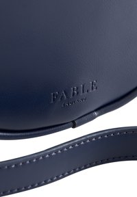 Sac bandoulière en cuir marine avec le logo embossé "FABLE ENGLAND". Il présente une texture lisse et une accentuation de la sangle cousue.
