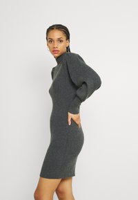Vestido de punto acanalado gris con cuello alto, mangas abullonadas y bolsillos laterales. La tela tiene una apariencia suave y texturizada.