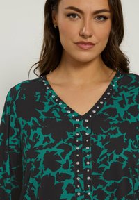 Zwarte en teal-gepatroneerde blouse met een V-hals, versierd met metalen studs langs de halslijn en knoopsluiting, met lange mouwen.