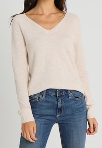 Pull beige en maille avec col en V et poignets côtelés, associé à un jean bleu. Le tissu présente un motif texturé et une coupe décontractée.
