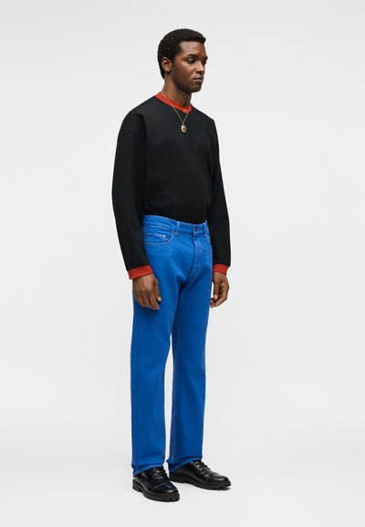 Άνδρας που φοράει μαύρο μακρυμάνικο πουκάμισο με κόκκινες μανσέτες και γιακά, φωτεινά μπλε παντελόνια, μαύρα παπούτσια και κολιέ με μενταγιόν, στέκεται σε λευκό φόντο.