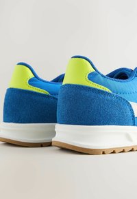 Baskets de course en daim bleu et synthétique avec une semelle intermédiaire blanche, présentant un accent jaune vif au niveau du talon et des détails de surface texturés.