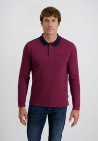 Langarm-Polohemd in tiefburgunder mit schwarzem Kragen, ausgestattet mit einer Brusttasche und ohne sichtbare Muster. Kombiniert mit dunkelblauen Jeans.
