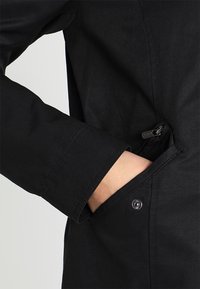 Černá bunda s kapsou na zip a detailním knoflíkem, vyrobená z hladké látky. Rukáv má přesný střih a čisté švy.