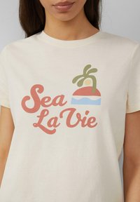Crème-kleurige katoenen t-shirt met de tekst "Sea La Vie" in roze en groen, met een grafisch ontwerp van een gestileerd strandbeeld boven de tekst.