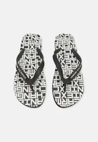 Svarte og hvite flip-flopper med et gjentagende tekstmønster og svarte gummistropper med "Armani Exchange"-merke. Flat såledesign.