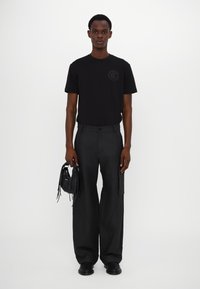 Camiseta de algodón negra con un logo redondo, combinada con pantalones anchos negros. El modelo sostiene un pequeño bolso de cuero negro con detalles de flecos.