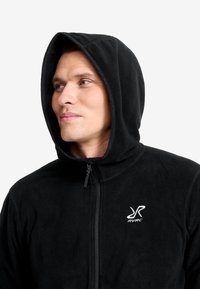 Schwarze Fleecejacke mit Reißverschluss vorne, Kapuze und aufgesticktem Logo auf der linken Brust. Weiche Textur, schlankes Design.