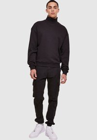Svart hettegenser med høy krage, kombinert med svarte cargo-bukser med flere lommer og hvite joggesko. Glatt tekstur, avslappet passform.