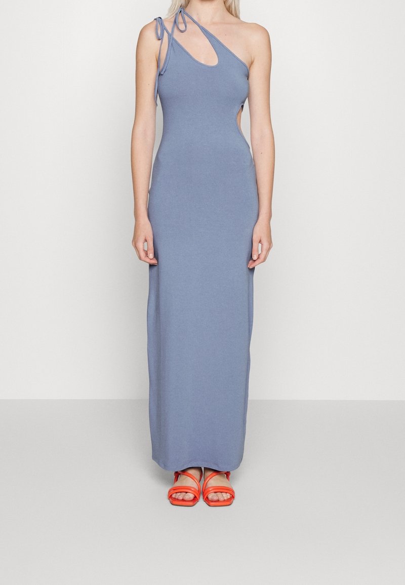 Femme portant une robe bleue, longue jusqu’aux chevilles, sans manches, à une épaule avec un détail découpé et des sandales orange, debout devant un fond uni.