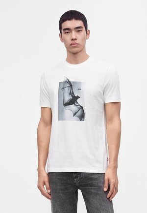 Jeune homme aux cheveux courts et foncés portant un t-shirt blanc avec un grand motif chauve-souris en noir et blanc et un jean gris foncé.