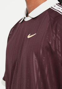 Polo desportivo cor bordô com colarinho branco, apresentando um corte slim, riscas verticais discretas e um logo dourado da Nike no lado esquerdo do peito.