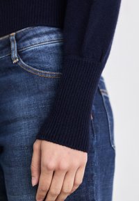 Navy-blauer Strickpullover mit gerippten Bündchen, kombiniert mit Jeans in mittlerer Waschung mit orangefarbener Naht und klassischem Taschen-Design.