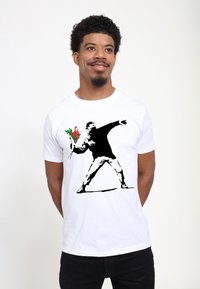 Bílý bavlněný tričko s černým grafickým motivem ve stylu šablony, zobrazující postavu házející květiny, s červenými a zelenými akcenty na kytici.