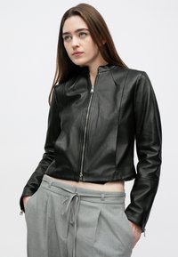 Casaco de couro preto cropped com fecho de correr frontal, design ajustado, textura suave e detalhes em costura, combinado com calças cinza claro.