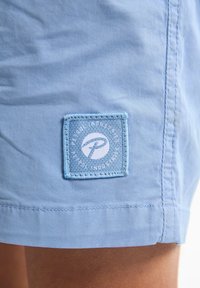 Ljusblå tygshorts med en fyrkantig logotypapplikation med text och en grafisk emblem. Slät textur med sydda fållar.